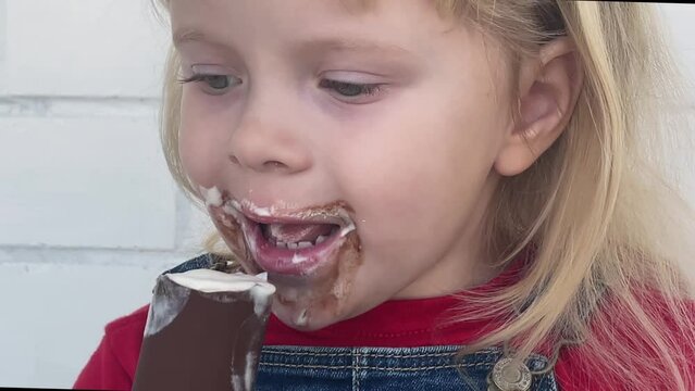 4k. Close up happy caucasian little girl eating ice cream. Her face is all smeared with ice cream.
