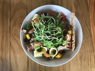 bowl of fresh green oak salad with sunflower sprouts, smoked bacon, dried tomatoes, steamed pumpkins, black olive, cheese and one roll cracker on brown wooden table