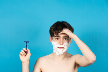 Worried shirtless teenager boy looking at camera while holding razor on his hands. Shaving fear on...
