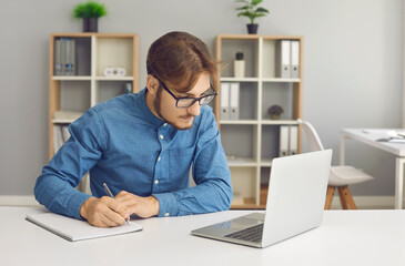 Fototapeta na wymiar Concentrated young man with glasses makes notes while working with laptop at home. Businessman or student jotting down information from internet or important things during distance business training.