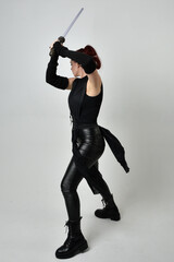 Obraz premium Full length portrait of pretty red haired female model wearing black futuristic scifi leather costume, holding a lightsaber sword weapon. Dynamic standing poses on a white studio background.