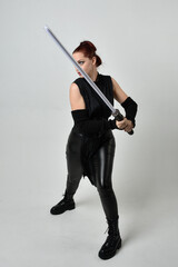 Full length portrait of pretty red haired female model wearing black futuristic scifi leather costume, holding a lightsaber sword weapon. Dynamic standing poses  on a white studio background.