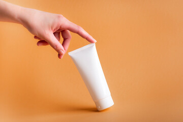 female hand touches a white tube of body cream on an orange background