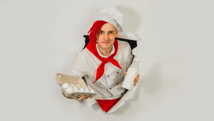 Man with red dreadlocks holds it in his hand chicken eggs and milk. Confident young cook in white apron and red tie holding chicken eggs and a bottle of milk, peeking through ripped paper background