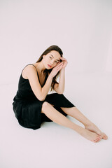 Young slim brunette woman in black dress sits barefoot on floor.