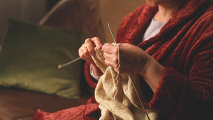 Unrecognizable senior female in warm bathrobe sitting on couch and knitting in dim living room in...