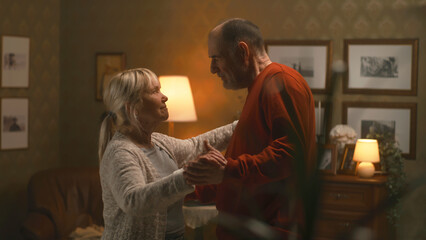 Aged spouses in love talking and dancing a romantic slow dance in the evening at home in a cozy living room with lamps and paintings