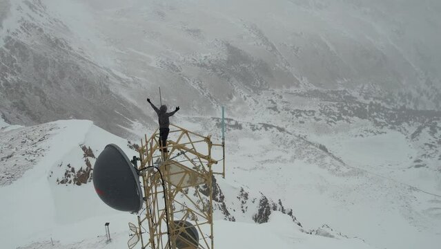 Aerial View of Man on Top of Communication Tower in Snow Capped Mountain Peak Raising Arms, Drone Shot