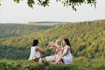 Company of young female friends having fun, drink wine, and enjoy hill landscape and summer picnic.