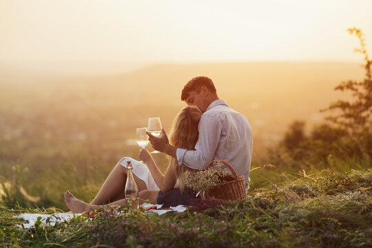 Attractive couple in love enjoying picnic on the hill at sunset. Happy young couple kissing and drinking wine. Romance, dating and love concept.