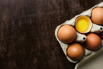 Fresh brown eggs and a broken egg with yolk in an eco tray made from recycled paper on a dark wooden background