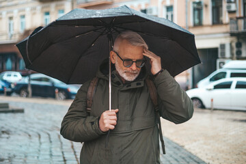 Mature man with migraine in rainy weather under an umbrella. Tiredness and headache when the...