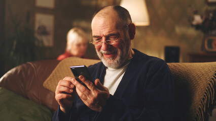 Cheerful elderly male in glasses chuckling and reading news while sitting on sofa, and browsing social media on smartphone on weekend day at home