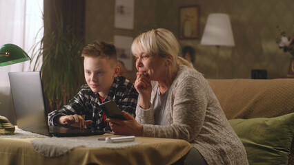 Teenager with elderly grandmother playing smartphone and laptop together at home sitting on the...