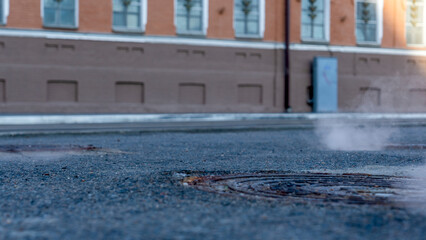 Manhole cover in a paved street on a cold day, as steam escapes from the sewer below up into the city street.