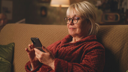 An elderly woman in a bathrobe texting and watching video on the phone while sitting on the sofa in...
