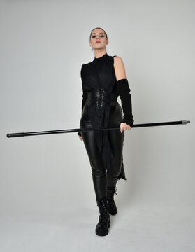 Full length portrait of pretty red haired female model wearing black futuristic scifi leather costume, holding a staff spear weapon. Dynamic standing pose on a white studio background.