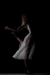 Girl with curly hair making ballet poses. Side lit silhouette of ballerina in white dress and black boots against black background.