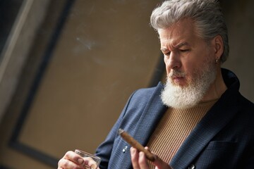 Close up portrait of serious mature man with beard looking at cigar while holding it and glass of...