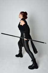 Full length portrait of pretty redhead female model wearing black futuristic scifi leather costume, holding a staff spear weapon. Dynamic standing pose facing away, backwards, white studio background.