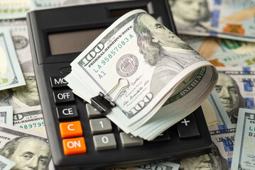 Close up photo of one hundred dollar bills with black binder clip on calculator on dollar banknotes background