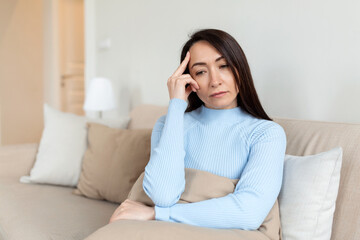 Frustrated Asian lady sitting on sofa, cuddling pillow, looking away at window. Lost in thoughts...