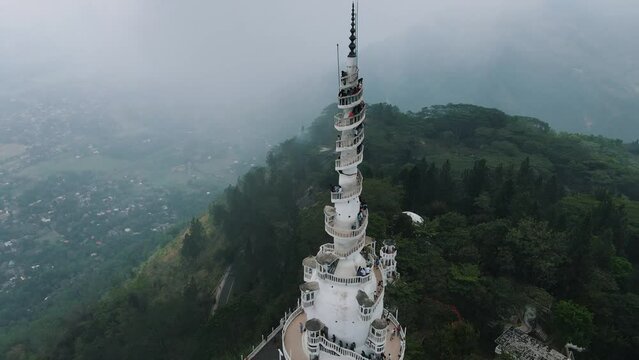 Beautiful high white Ambuluwawa tower between the green nature where several tourists at the top of the spiraling stairs enjoy the view of Sri-Lanka on a misty day. Backwards drone tilt shot
