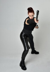 Full length portrait of pretty red haired female model wearing black futuristic scifi leather costume, holding a staff spear weapon. Dynamic standing pose on a white studio background.