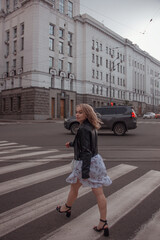 young curly blonde girl in white dress and black leather jacket is walking along the crosswalk and looking away in step on the white city buildings background at sunset. lifestyle concept, free space