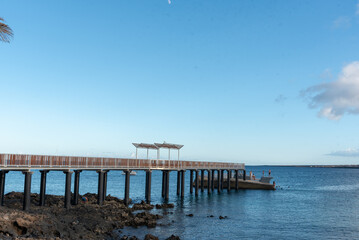 Fototapeta na wymiar Paisaje costero en Lanzarote Islas Canarias, Muelle la Garita en Arrieta que se adentra en el mar turquesa durante un día de verano soleado y con el cielo despejado. Recursos turísticos naturales.