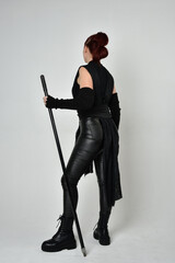 Full length portrait of pretty redhead female model wearing black futuristic scifi leather costume, holding a staff spear weapon. Dynamic standing pose facing away, backwards, white studio background.