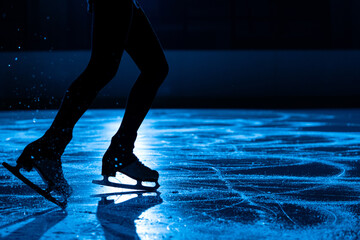 Dark silhouette of female legs in figure skating skates sliding on ice arena. Young woman trains on...