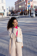 Happy curly brunette girl speaking on phone outdoors. Young woman happy walking in street. Pink sweater, beige coat, beige hat. Sun in city. Fashionable asian girl with frizzly hair with iphone