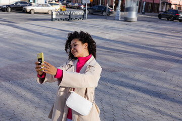 Happy curly brunette girl doing selfie on phone outdoors. Young woman happy walking in street. Pink sweater, beige coat, beige hat. Sun in city. Fashionable asian girl with frizzly hair with iphone