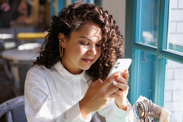 	
serious curly girl working in cafe with iphone. Woman typing on her phone. asian caucasian pretty young woman working in cafe