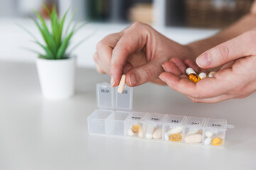 Female elderly hands sorting pills. Closeup of medical pill box with doses of tablets for daily take a medicine with white, yellow drugs and capsules. Young woman getting her daily vitamins at home.  - obrazy, fototapety, plakaty
