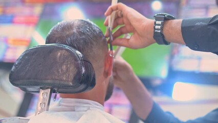 Back view of a man watching a football match during barber cuts his beard with a straight-edge razor in a barbershop. 