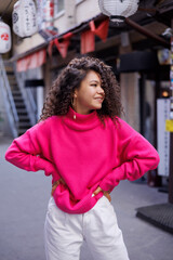 Happy curly brunette girl in sun glasses and hat smiling outdoors. Young woman happy walking in street. Pink sweater, beige coat, beige hat. Sun in city. Fashionable asian girl with frizzly hair
