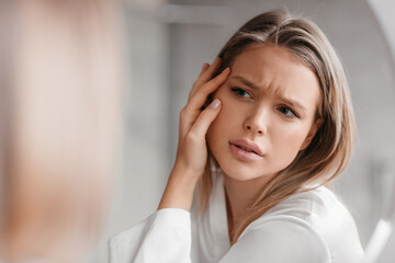 Acne prone skin concept. Sad lady looking at mirror and checking her face skin, standing in...
