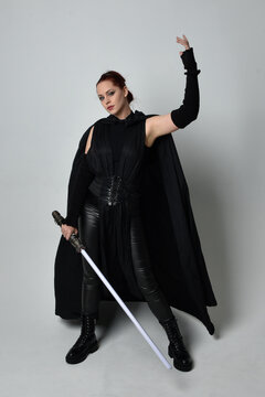 Full length portrait of pretty redhead female model wearing black futuristic scifi leather cloak costume, holding a lightsaber weapon. Dynamic standing pose on white studio background.