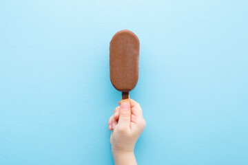 Baby boy hand holding ice cream with chocolate glaze on light pastel blue table background. Closeup. Children cold sweet snack in summer. Top down view.