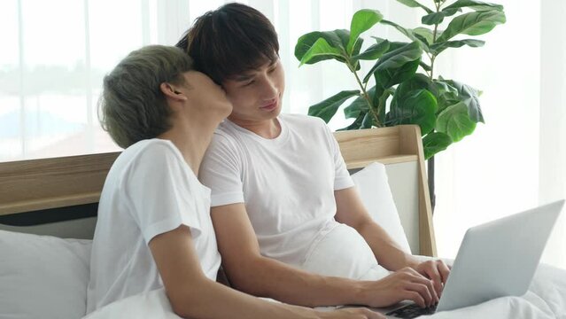 Indoors, medium shot portrait of young Asian gay couple teasing, kissing eac other, staying in bed, working in the morning, using laptop computer. Busy LGBT lifestyle, romantic concept.