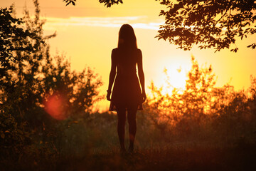 Back view of young woman in summer dress walking alone through evening dark forest