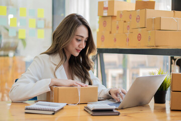 Smiling Asian woman looks at boxes and uses laptop to check information in parcel delivery boxes before sending them to customers. Entrepreneurs, small businesses work at home work online marketing