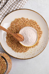 Buckwheat flour in a ceramic bowl with spoon and raw green buckwheat grain on a ceramic plate on light grey concrete background, top view. Alternative flour, gluten free flour, healthy nutrition