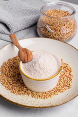 Buckwheat flour in a ceramic bowl with spoon and raw green buckwheat grain on a plate with napkin on light grey concrete background, close up. Alternative flour, gluten free flour, healthy nutrition