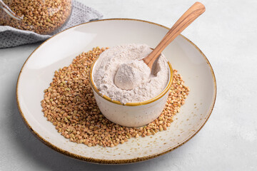 Buckwheat flour in a ceramic bowl with wooden spoon and raw green buckwheat grain on a plate on light grey concrete background, close up. Alternative flour, gluten free flour, healthy nutrition