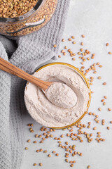 Buckwheat flour in a ceramic bowl with wooden spoon and raw green buckwheat grain in a jar on light grey concrete background, close up. Alternative flour, gluten free flour, healthy nutrition