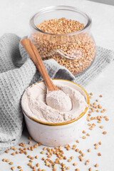 Buckwheat flour in a ceramic bowl with wooden spoon and raw green buckwheat grain in a jar on light grey concrete background, close up. Alternative flour, gluten free flour, healthy nutrition