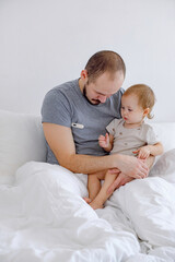 Fototapeta na wymiar Baby and man sitting on the bed on white linen. Toddler looking at father taking temperature.